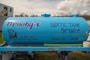 A blue septic tank service truck with "Orooby's Septic Tank Service" and humorous slogan written on it, parked outdoors.