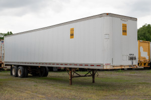 A white A-Verdi storage trailer with yellow signage, stationed on a grassy field with other containers and trailers in the background, under cloudy skies.