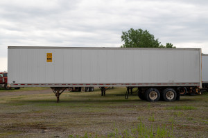 A white freight trailer with A-Verdi signage, parked on an empty lot with sparse vegetation and overcast skies.