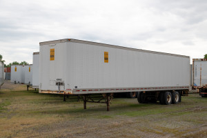 A white trailer with A-Verdi branding, parked on a grassy lot with other trailers and containers in the background, under cloudy skies.