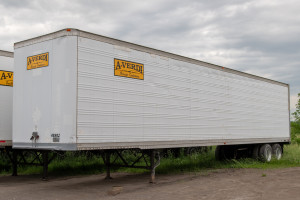 A white A-Verdi storage container trailer parked on a dirt ground with greenery in the background under a cloudy sky.