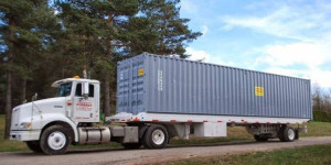 A white semi-truck hauling a large gray shipping container on a flatbed trailer, parked on the side of a road with trees in the background.