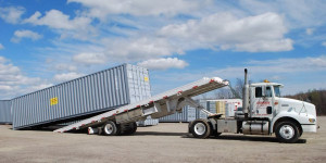 A white semi-truck with a tilting flatbed trailer unloading a large gray shipping container in an open area with a clear sky above.